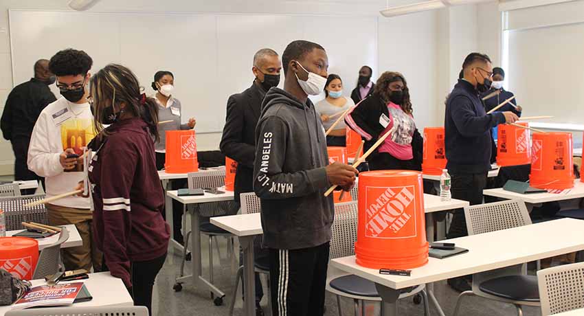Students use drumsticks to pound on inverted orange utility buckets
