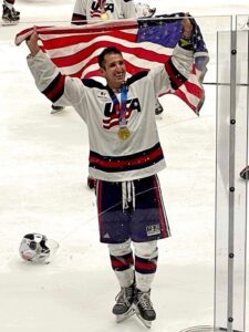 Max Roth stands on ice in his hockey uniform, holding the American flag above his head