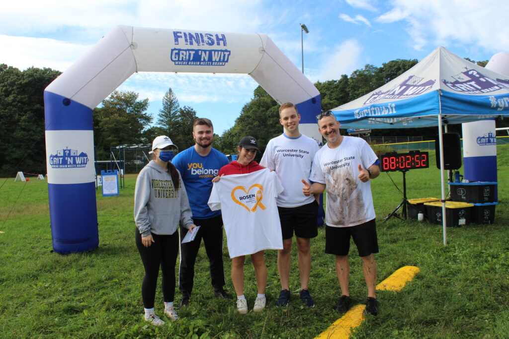 Five students pose in front of the finish line of the course. The student in the center holds up a T-shirt with Rosen Cancer Awareness Fund printed on it.
