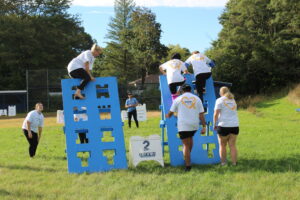 Three students climb a seven-foot-high obstacle that looks like a blue board with large letters cut into it for hand- and footholds.