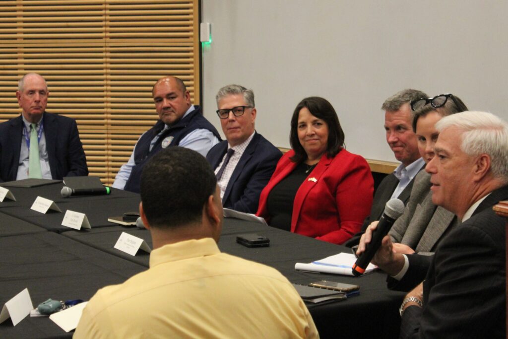 A group of people in business attire sit around a conference table, engaged in a discussion, with one man speaking into a microphone.