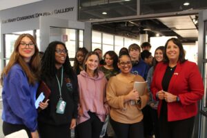A group of students and an adult stand together smiling in front of the entrance to The Mary Cosgrove Dolphin Gallery. Some hold notebooks and wear name badges.