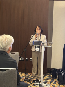 A woman in business attire stands at a podium, speaking into a microphone at an indoor event. An audience member is seated in the foreground.