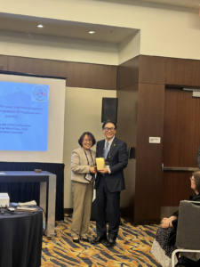 Two people standing side by side in a conference room, smiling and holding a yellow object, with a projection screen and audience members visible in the background.
