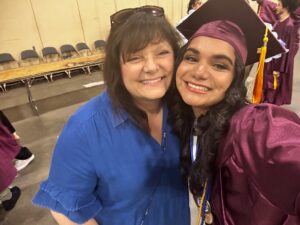 A graduate in a maroon cap and gown smiles beside a woman in a blue shirt; they are indoors with chairs and a table in the background.