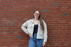 A woman with long hair, glasses, and a light jacket stands smiling in front of a red brick wall.
