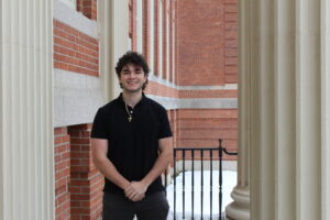 A young man in a black shirt stands smiling between brick and columned buildings in an outdoor walkway with snow on the ground.