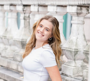 A young woman in a white crop top and blue jeans stands against a stone balustrade, smiling with her hands in her pockets.
