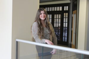 A woman with long hair stands indoors by a railing, smiling, with glass doors and windows in the background.