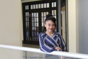 A woman in a blue and white striped blouse stands indoors, leaning on a white railing, with dark wooden doors in the background.