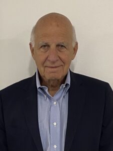 An older man with a bald head, wearing a dark suit jacket and light blue shirt, stands in front of a plain light-colored wall.