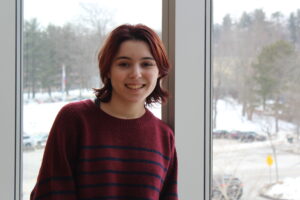 A person with short, dark reddish-brown hair and a striped maroon sweater stands by a window with a snowy scene and trees visible outside.