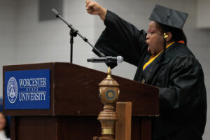 A graduate in cap and gown raises a fist while speaking at a podium labeled "Worcester State University" during a commencement ceremony.