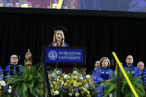 A speaker in graduation attire stands at a podium labeled "Worcester State University," addressing an audience at a commencement ceremony. Faculty in blue robes are seated behind her.