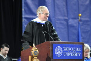 A speaker in academic regalia stands at a podium labeled "Worcester State University" during a formal event.