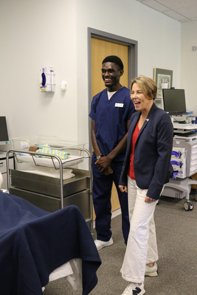 A nurse and a woman in business attire stand and smile in a hospital room next to an incubator with a baby doll inside.