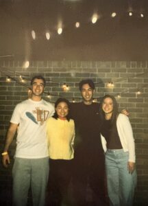 Four people stand close together and smile in front of a brick wall with string lights at night.