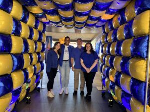 Four people stand together inside a tunnel decorated with alternating blue and yellow balloons, smiling at the camera in a brightly lit indoor space.