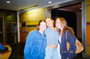 Three young women stand close together and smile for the camera in a brightly lit indoor setting.