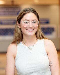A young woman with long hair and clear-framed glasses smiling, wearing a white tank top and a cross necklace.
