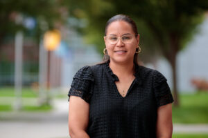 A woman wearing glasses and a black top smiles while standing outdoors.