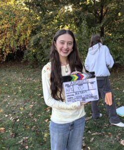 A smiling woman holding a film slate labeled “Anomalia” in front of trees, with another person standing in the background.