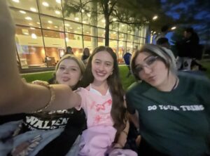 Three young women taking a selfie outdoors in front of a glass building at night.