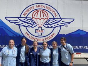 Six medical professionals in blue scrubs stand in front of a Remote Area Medical Volunteer Corps vehicle.