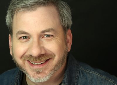 A close shot of Steve Gagliastro smiling into the camera in front of a black background. He has short hair and a beard and mustache and is wearing a dark denim shirt.