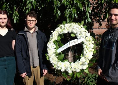 Renee Mercier '22, Preston Carr '22, and Daniel Bourget '25 stand next to a memorial wreath of white flowers with a ribbon that says, 