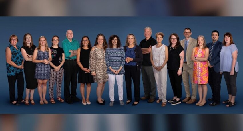 16 faculty stand in a row in front of a blue background