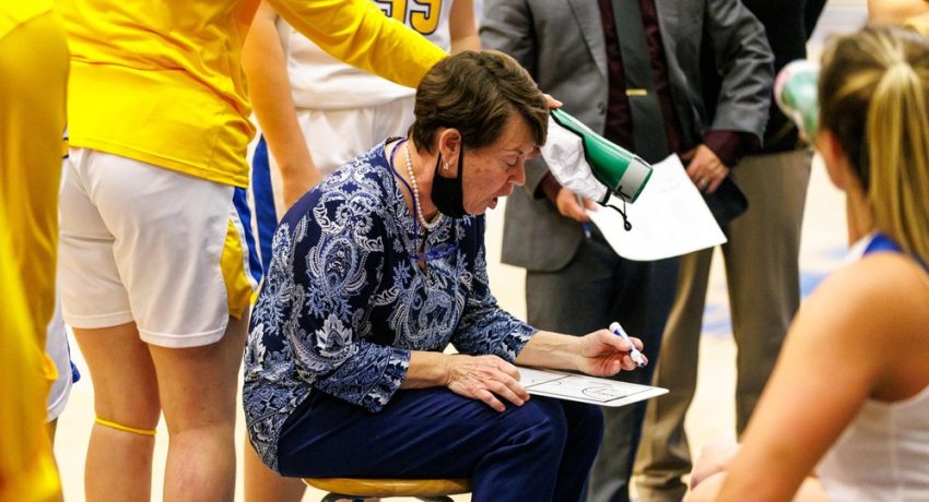 Basketball coach seated on the sidelines at a game writing on a board with players around her writing on