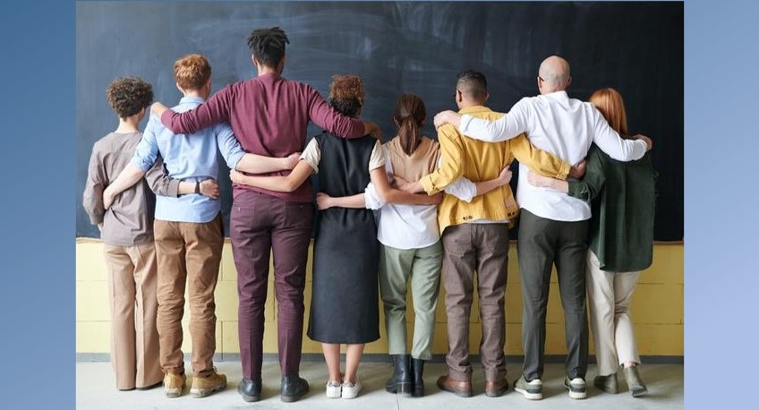 8 people of varying heights, ethnicities, and genders stand in a line facing a chalkboard, their backs to us and their arms around one another