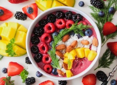 bowl with yogurt, blackberries, blueberries, mango, coconut and almonds sitting on a surface surrounded by more fruit