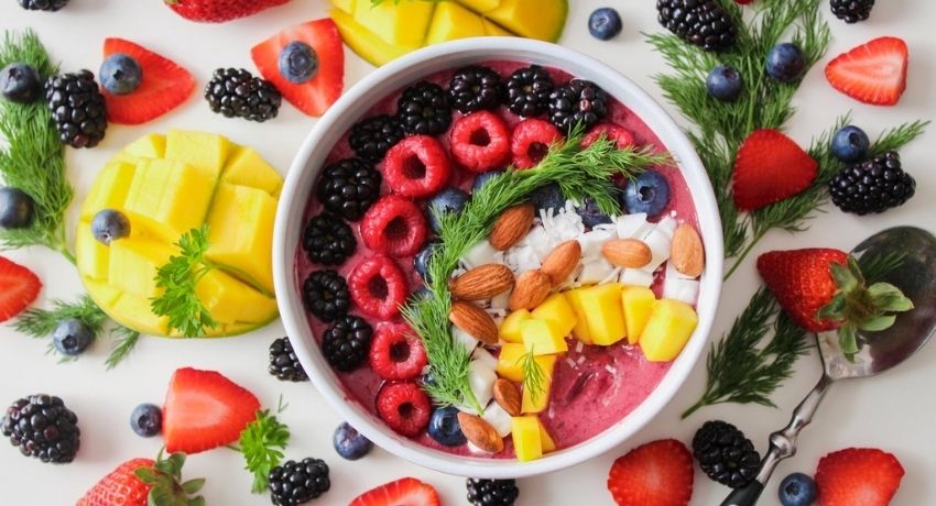 bowl with yogurt, blackberries, blueberries, mango, coconut and almonds sitting on a surface surrounded by more fruit