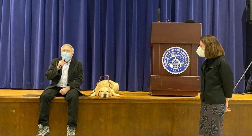 man in mask sits on stage with a microphone, dog beside him as female professor looks on