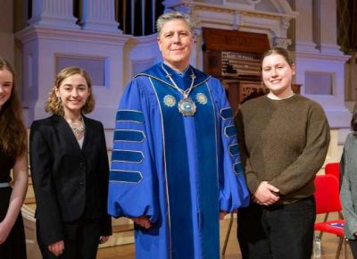 University president in academic robes poses on stage with four female students