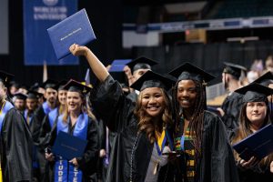 Students in gap and gown smile and one holds up her Worcester State University degree