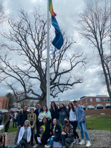 Eleven students from the LGBTQ+ Narratives class and Professor Riley McGuire pose at the base of the flagpole with the new Pride flag and Hate Has No Home Here flag