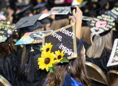 Rows of seated Graduating college students in caps and gowns with one cap reading teach, love inspire