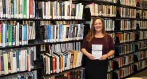 Professor Aimee Delaney stands next to shelves of books in the Worcester State library, holding a copy of her own book