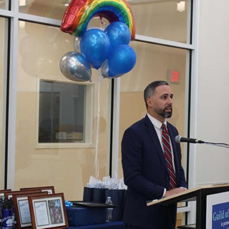 Man in suit at podium with blue and rainbow balloons behind him