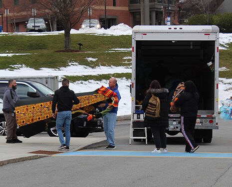 Three people lift a large doll from a truck while three other people remove items from back of truck