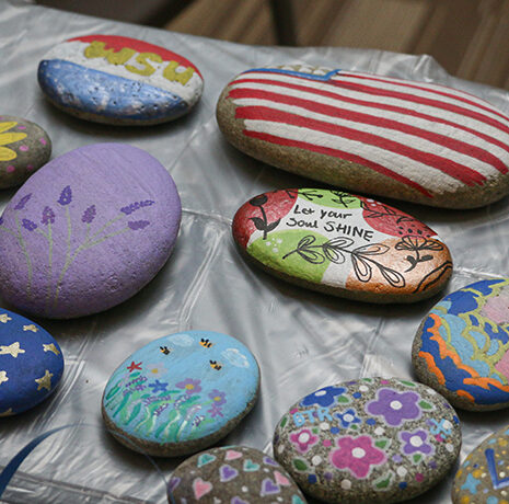 Several smooth stones are decorated with colorful painted designs, patterns, and messages, displayed on a table covered with a plastic sheet.