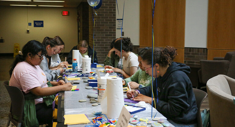 A group of people sit around a table painting rocks and crafting in an indoor setting with supplies and decorations on the table.
