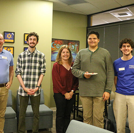 Five people stand side by side in an office lounge area, posing for a group photo. Some are smiling, and one person is holding a small object.