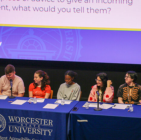 Six panelists sit at a blue table on a stage under a screen displaying a discussion question at a Worcester State University event.