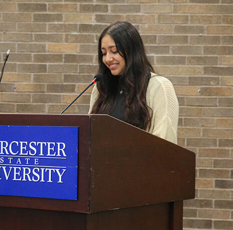 A woman stands at a podium with a Worcester State University sign, speaking into a microphone in front of a brick wall.