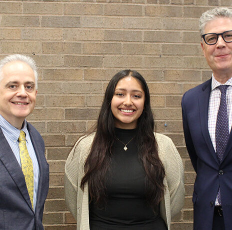 Three people, two men in suits and one woman in a black top and light cardigan, stand in front of a brick wall, facing the camera and smiling.