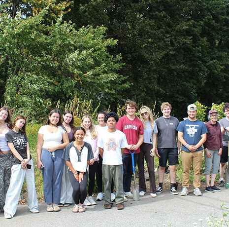 A group of sixteen young people stand in a row outdoors on a path, surrounded by greenery, looking at the camera.